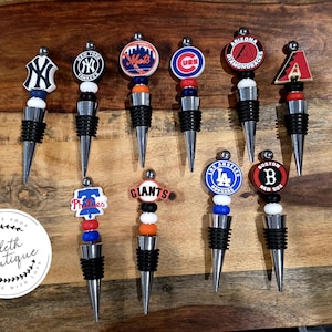 May include: A collection of baseball team logo bottle stoppers. Each stopper features a team logo, with a black rubber base and a silver metal spike. The logos include the Yankees, Mets, Cubs, and more.