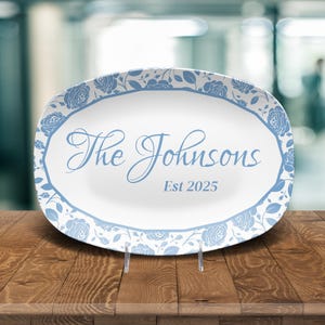 Puede incluir: Plato ovalado con centro blanco y borde floral azul. El plato presenta el texto "The Johnsons" y "Est 2025" en escritura azul claro. El plato se exhibe sobre una superficie de madera.