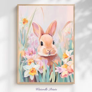 Puede incluir: Una impresión artística enmarcada que presenta un conejo marrón claro rodeado de flores coloridas y follaje verde. El conejo tiene ojos grandes y oscuros y orejas largas. Las flores son blancas, amarillas y rosas. La impresión tiene una paleta de colores pastel.