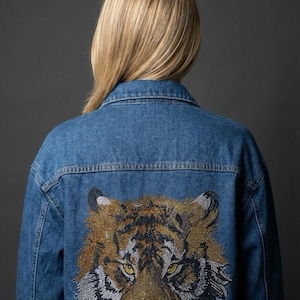 May include: A denim jacket with a detailed tiger face design on the back. The tiger is rendered in shades of gold, black, and silver, with yellow eyes. The jacket is a classic blue wash.