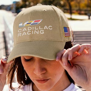 May include: Tan baseball cap with the text "CADILLAC RACING" embroidered in white. The cap features a colorful logo and a small American flag patch on the side. The cap is made of a durable fabric and has a curved brim.