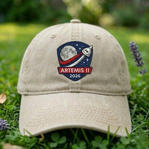 May include: A tan baseball cap featuring an embroidered patch with a moon, rocket, and the text "ARTEMIS II 2026". The cap is made of a soft fabric and has a curved brim. The background is blurred green grass.