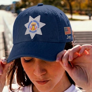 May include: Navy blue baseball cap with a white and gold Private Security Officer patch. The cap features an American flag patch and the name "Jay" embroidered on the side. A classic, adjustable hat.