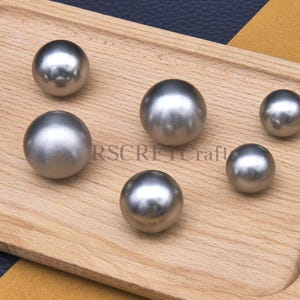 Op de afbeelding: Een houten dienblad met zeven glanzende, zilverkleurige metalen bollen van verschillende grootte. De bollen liggen op het dienblad, dat tegen een achtergrond van marineblauw en mosterdgeel staat. De tekst "RSCRET Craft" is zichtbaar op het dienblad.