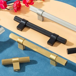 May include: An assortment of cabinet pulls in various metallic finishes, including silver, black, and gold. Each pull features a ribbed, cylindrical handle and a square base. The pulls are displayed on a neutral background with floral accents.