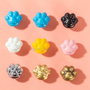 May include: Nine paw-shaped cabinet knobs in various colors and finishes, including blue, black, white, yellow, pink, silver, gold, and bronze. The knobs are arranged in a grid pattern against a pink background. These decorative knobs are ideal for pet lovers.