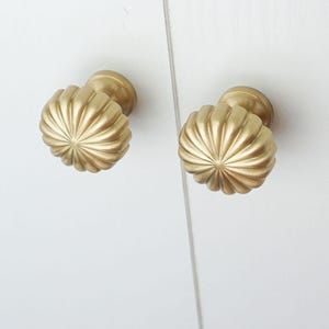 May include: Two gold cabinet knobs with a fluted, rounded design. The knobs are mounted on a white surface, suggesting furniture or cabinetry. The hardware has a classic, decorative style.