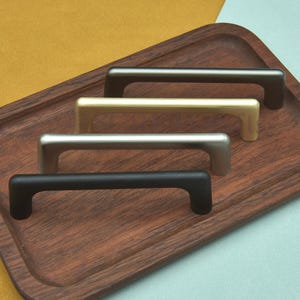 May include: Four cabinet pulls in different metallic finishes: black, silver, gold, and bronze. The pulls are arranged on a wooden tray with a dark brown finish. The background is a split of yellow and light blue.