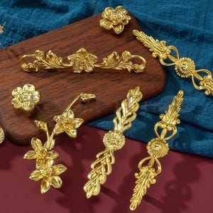 Puede incluir: Una colección de herrajes dorados y ornamentados para armarios, que incluyen pomos y tiradores. Los herrajes presentan diseños florales y de hojas, con diferentes formas y tamaños. Las piezas están dispuestas sobre una tabla de madera sobre un fondo azul y granate.