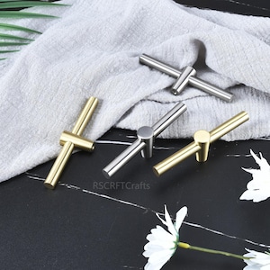 May include: A collection of cabinet pulls in gold and silver tones. The pulls have a T-bar design with a cylindrical base. The hardware is displayed on a black surface with a gray cloth and white flowers.