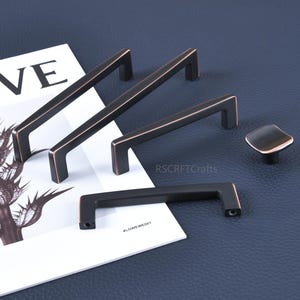 May include: A collection of bronze-colored cabinet hardware, including several rectangular bar pulls and a square knob. The hardware is displayed on a white paper with the word "WE" visible, set against a dark blue background.