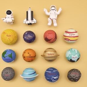 May include: A collection of miniature space-themed figurines and planets. Includes an astronaut, a shuttle, and planets like Earth, Mars, and Saturn. The planets are colorful and labeled, set against a yellow background.