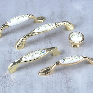 May include: A collection of cabinet hardware featuring gold-toned handles and a knob. The handles have a white ceramic center with a floral design in blue, pink, and green. The hardware is designed for furniture.