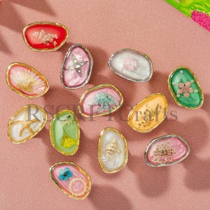 May include: An assortment of oval resin pendants with gold-coloured edges. Each pendant showcases a unique design, such as dried flowers, starfish, and seashells, set against diverse coloured backgrounds. The pendants are displayed on a pink surface.