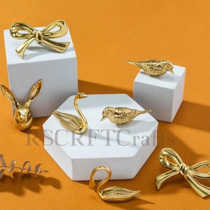 May include: A selection of gold-coloured decorative items, including a bow, a rabbit head, swans, and birds. The items are displayed on white geometric platforms against an orange background. These are likely for home decor.