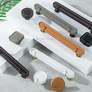 May include: A collection of leather cabinet pulls and knobs in various colors, including gray, brown, tan, and white. The pulls have a stitched design and are paired with matching knobs. The hardware is displayed on a white surface.