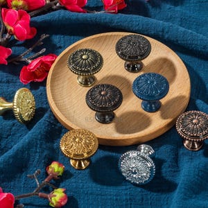 May include: A collection of decorative cabinet knobs in various metallic finishes, including gold, bronze, and silver, with intricate embossed designs. The knobs are displayed on a wooden tray and a blue fabric background, with red flowers.