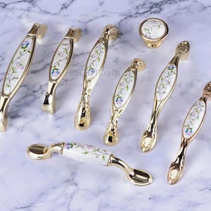 Op de afbeelding: Een verzameling goudkleurige meubelbeslag, waaronder handgrepen en een knop. Elk stuk heeft een witte keramische inleg met een delicaat bloemmotief in blauw, paars en groen. Het beslag is geplaatst op een marmeren oppervlak.