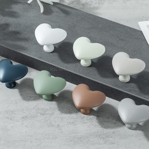 Peut inclure: Ensemble de boutons de meubles en forme de cœur, de couleurs variées : blanc, bleu, vert et marron. Les boutons sont disposés sur une surface grise, mettant en valeur leur design et leurs variations de couleurs. Idéal pour ajouter une touche de charme aux meubles.