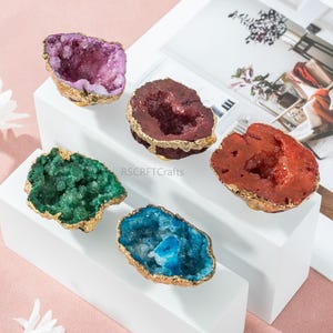 May include: Five colorful geode specimens with gold-painted edges are displayed on a white platform. The geodes are in shades of purple, burgundy, green, blue, and red. The image is set against a soft pink background.