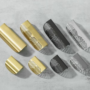 May include: A selection of cabinet pulls in gold, black, and silver finishes. Some have a geometric leaf design, others are plain. The hardware is displayed on a light grey surface. Measurements are in centimetres.