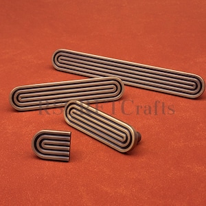 May include: A set of four bronze-toned cabinet pulls and knobs. The hardware features a grooved, rectangular design with a U-shaped pattern. The pulls and knobs are arranged on a reddish-orange background.