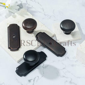 Puede incluir: Un surtido de pomos y placas traseras en varios acabados, incluyendo bronce y negro. Los herrajes se exhiben sobre bloques de piedra blanca sobre un fondo de mármol. El texto "RSCH Crafts" es parcialmente visible.