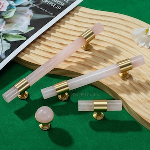 May include: A collection of rose quartz cabinet hardware, including two long pulls, a short pull, and a round knob. The hardware features gold-toned metal accents. The items are displayed on a wooden surface with a green background. The text "CREATIVE MULTIPURPOSE PORTFOLIO" is visible.
