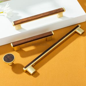 May include: Cabinet hardware set with a dark wood and gold finish. The set includes two rectangular handles, a longer bar handle, and a round knob. The hardware is displayed on a white surface against a yellow background.