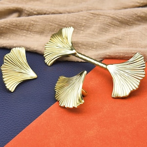 May include: Gold-colored ginkgo leaf-shaped cabinet hardware set. The set includes two knobs and a pull, all with a detailed, textured design. The hardware is displayed on a blue and orange surface with a tan fabric background.
