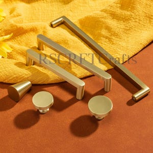 Könnte beinhalten: Eine Sammlung von Messing-Schrankbeschlägen, darunter Griffe und Knöpfe, angeordnet auf einem gelben Tuch. Das Set umfasst verschiedene Größen und Formen, mit einem kegelförmigen Knopf und mehreren runden Knöpfen. Der Text "RSCRFTCrafts" ist sichtbar.