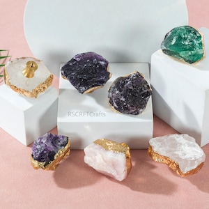 May include: A selection of gemstone cabinet knobs in shades of purple, green, and white. Each knob features a gold-coloured trim and is presented on a white plinth against a pink backdrop. The text "RSCRFTCrafts" is visible.