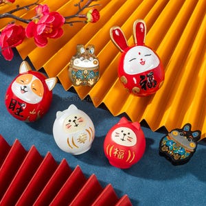 Pode incluir: Uma coleção de figuras decorativas coloridas, incluindo coelhos, gatos e um cão, com caracteres chineses. As figuras são vermelhas, brancas e pretas, com detalhes dourados. O fundo inclui um leque amarelo e flores vermelhas.