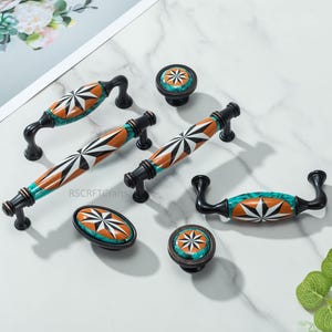 以下が含まれることがあります： 装飾的なキャビネットハードウェアのコレクション。ハンドルとノブが含まれています。オレンジ、白、黒のスターバーストデザインで、ターコイズと黒のアクセントが付いています。ハードウェアはセラミックと金属製です。