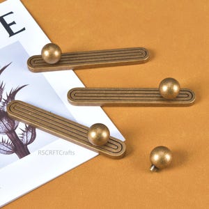 May include: Antique brass cabinet pulls with a ball-shaped knob. The pulls have a rectangular base with a grooved design. One knob is detached. The pulls are on a tan surface with a magazine.