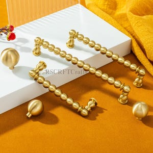 Puede incluir: Juego de herrajes dorados para armarios, que incluye dos tiradores de barra con cuentas y dos pomos redondos. Los herrajes se muestran sobre una superficie blanca sobre un fondo color mostaza. El texto "RSCRFTCraft" es visible.