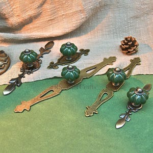 May include: A selection of antique cabinet handles and knobs. The hardware features green ceramic pumpkin-shaped knobs on ornate bronze-coloured metal backplates and pulls. A pine cone rests nearby on a green surface.