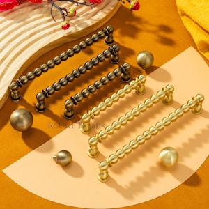 Op de afbeelding: Een assortiment decoratieve kastbeslag in antiek messing en goudtinten. De collectie omvat verschillende kralen handgrepen en bijpassende ronde knoppen. De hardware wordt gepresenteerd op een warme, neutrale achtergrond met een houten accent en bloemstuk.