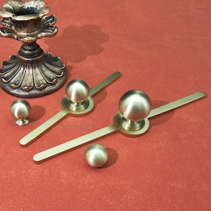 May include: An assortment of antique brass hardware, including drawer pulls and a candlestick holder. The drawer pulls have a circular base with a long, thin handle and a spherical knob. The candlestick holder has a floral design.