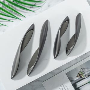 Może przedstawiać: Cztery nowoczesne, ciemnoszare uchwyty do szafek ułożone na białej powierzchni. Uchwyty mają zakrzywiony, wydłużony kształt z kontrastującym srebrnym wnętrzem. Uchwyty są w różnych rozmiarach, do różnych szuflad lub drzwi.