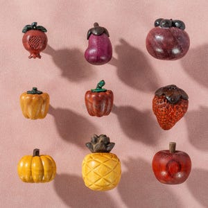 Op de afbeelding: Een verzameling van negen houten fruit- en groentefiguren. De collectie omvat een granaatappel, aubergine, mangosteen, paprika, aardbei, pompoen, ananas en appel. Elk stuk is ingewikkeld gesneden en beschilderd in realistische kleuren.