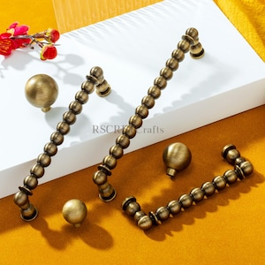 May include: Antique brass cabinet hardware set. Includes two long beaded handles, two small beaded handles, and two round knobs. The hardware is displayed on a white surface with a yellow background.