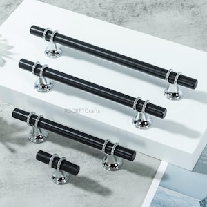 May include: A set of four black cabinet pulls with silver-colored accents. The pulls have a cylindrical design with a central bar and decorative rings. The base is a polished silver color. The brand name "RSCRFTCrafts" is visible.