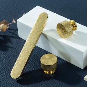 May include: A set of three brass hardware pieces, including a long, rectangular pull and two circular knobs. The hardware has a textured, ridged surface and is displayed on a white block against a dark blue background.