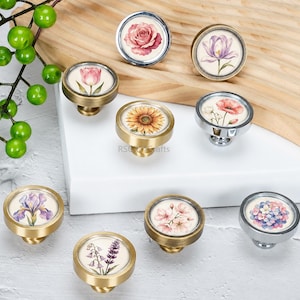 May include: A collection of nine round cabinet knobs featuring various floral illustrations including a rose, tulip, sunflower, poppy, iris, cherry blossoms, and hydrangea. The knobs have metallic finishes in gold and silver tones.