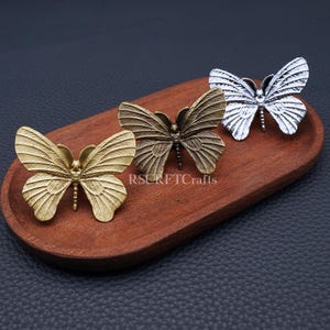 May include: Three metal butterfly ornaments in gold, bronze, and silver sit on a wooden tray. The butterflies have detailed wing patterns and are arranged in a row. The tray is oval-shaped and made of dark wood. The text "RSCRFTCrafts" is visible on the tray.