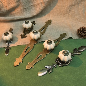 May include: A collection of antique-style cabinet pulls and knobs. The knobs are white with a dark center, and the pulls have ornate metalwork in bronze and dark silver tones. A pinecone is also present.