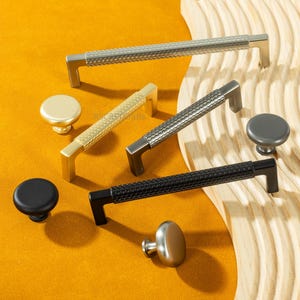 May include: An assortment of cabinet hardware, including silver, gold, gray, and black handles and knobs. The handles have textured patterns, and the knobs are round. The hardware is displayed on a yellow surface with a wooden accent.