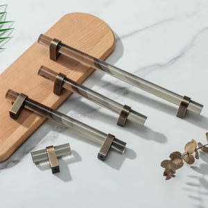 Peut inclure: Ensemble de quatre poignées de meubles avec des barres en acrylique transparent et des fixations en métal couleur laiton antique. Les poignées sont disposées sur une planche en bois et une surface en marbre blanc. La plus longue mesure environ 30 cm.