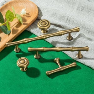 May include: A selection of antique brass cabinet hardware, including knobs and handles. The hardware features a bamboo-inspired design with a warm, aged finish. The pieces are arranged on a green surface with a wooden tray and fabric in the background.